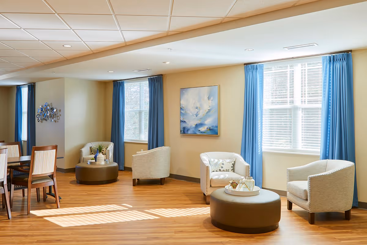 A bright and spacious living room area with wooden flooring, beige walls, and large windows with blue curtains. The room features comfortable beige armchairs arranged around round brown ottomans, a dining table with chairs, and decorative wall art. Sunlight streams through the windows, creating a warm and inviting atmosphere.