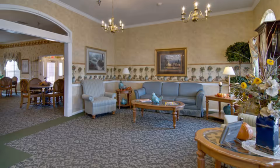 A cozy senior living facility common area with a blue sofa, a striped armchair, wooden coffee and side tables, decorative lamps, framed paintings on the wall, and floral wallpaper border. Adjacent dining area with wooden chairs and tables is visible through a large archway.