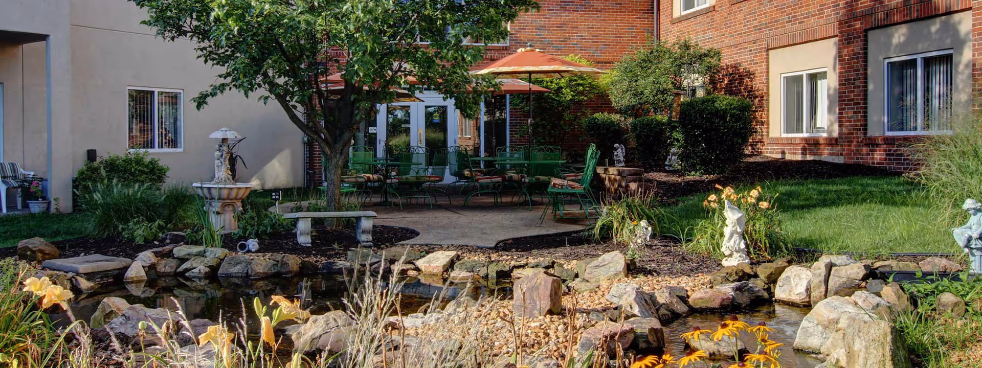 Outdoor courtyard area at Richmond Terrace featuring a small pond with rocks and flowers surrounding it, a stone bench, a fountain, green patio chairs and tables with an orange umbrella, and a brick building with windows in the background.