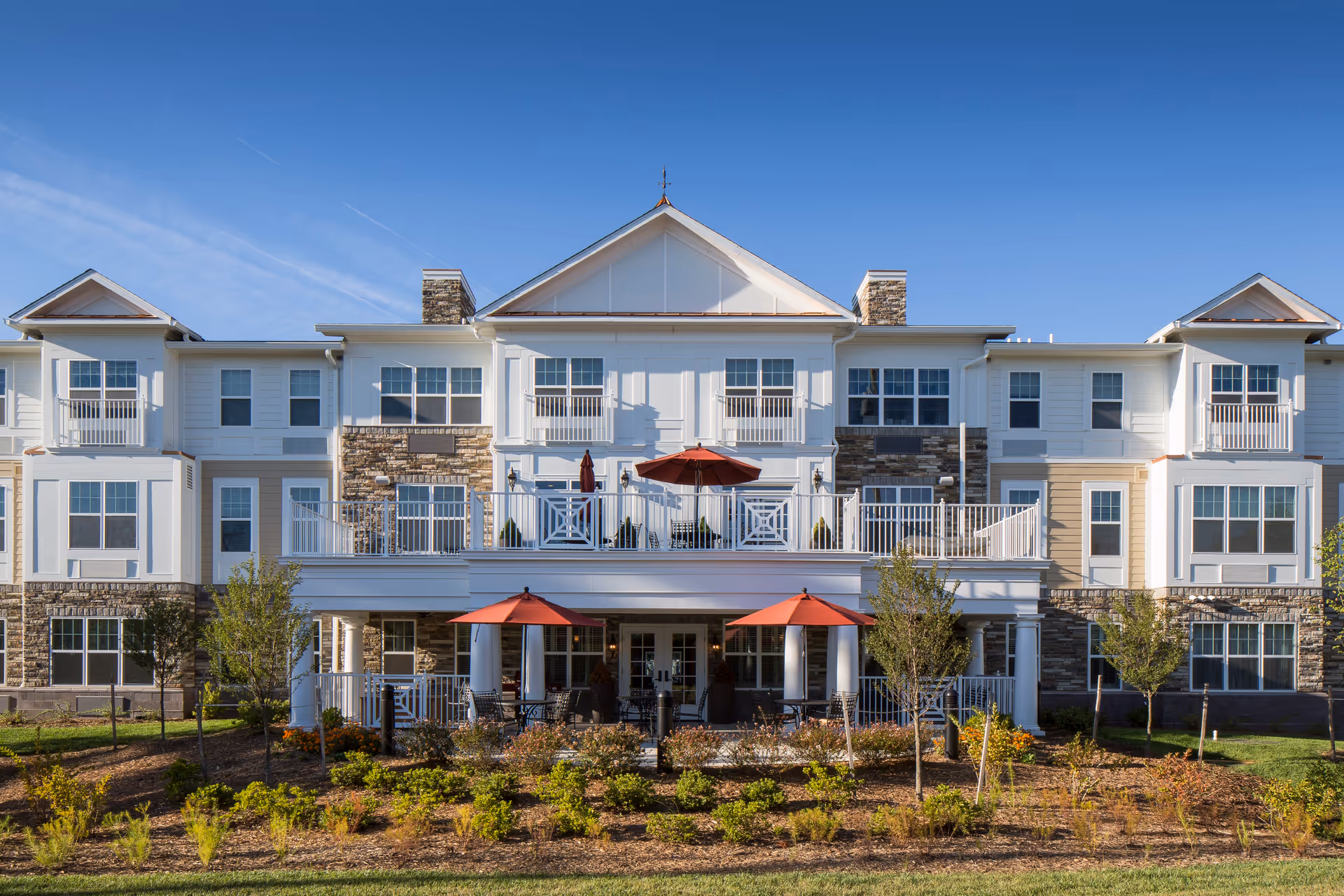 Exterior view of a multi-story senior living facility building with white siding and stone accents. The building features balconies and a patio area with red umbrellas and outdoor seating. There are small trees and landscaped bushes in front of the building under a clear blue sky.
