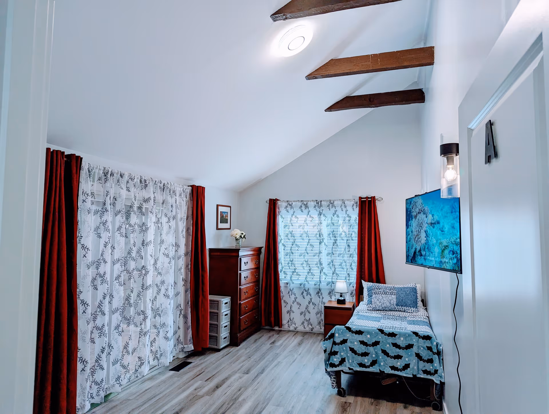 A bright bedroom with a single bed covered in blue and black patterned bedding. The room has light wood flooring, white walls, and a vaulted ceiling with exposed wooden beams. There are two windows with white sheer curtains and red drapes. A wooden dresser and a small nightstand with a lamp are also visible. A flat-screen TV is mounted on the wall opposite the bed.