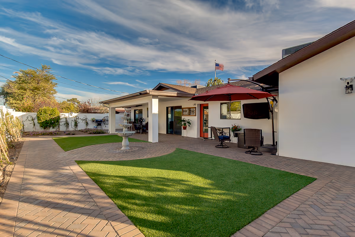 Outdoor patio area of West Drive Assisted Living and Memory Care featuring a paved walkway, green artificial grass patches, a white multi-tiered fountain, patio furniture with a red umbrella, and an American flag flying in the background under a partly cloudy sky.