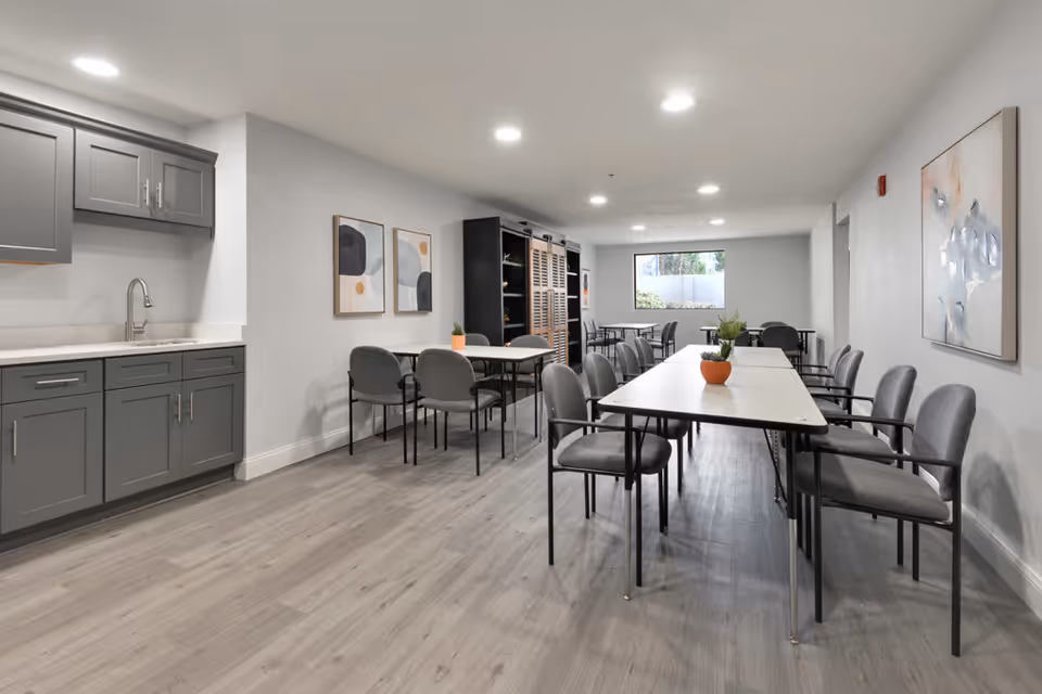 A modern dining area with multiple tables and gray cushioned chairs arranged neatly. The room features light gray walls, wood-look flooring, and recessed ceiling lights. On the left side, there is a kitchenette with gray cabinets and a sink. Abstract artwork is hung on the walls, and small potted plants decorate the tables. A window at the far end lets in natural light.