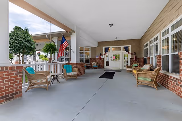 Covered entry porch with wicker chairs and tables, brick half-walls, potted plants, American flags, and double glass doors.