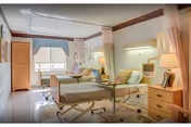 A bright and clean hospital-style room with two adjustable beds, each equipped with side rails and overbed tables. The room features soft lighting from wall-mounted lamps, a window with curtains, a wooden wardrobe, and a small bedside table with a lamp and personal items. The walls are light-colored with a decorative border near the ceiling.