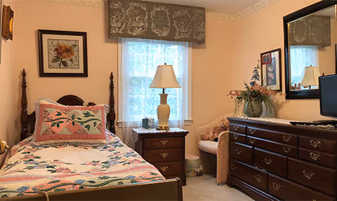A cozy bedroom with a single wooden bed featuring a floral quilt and matching pillow. Next to the bed is a wooden nightstand with a lamp and a small clock. A window with a decorative valance lets in natural light. Across from the bed is a large wooden dresser with a mirror above it, a vase with flowers, and a teddy bear sitting on a cushioned chair beside the dresser. The walls are painted a soft peach color and decorated with framed artwork.