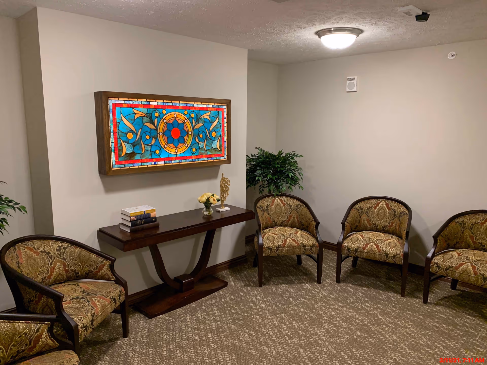 A small seating area in a retirement community with four patterned armchairs arranged around the walls, a wooden console table with books and a small flower arrangement, a colorful stained glass artwork mounted on the wall above the table, and a potted plant in the corner under a ceiling light.