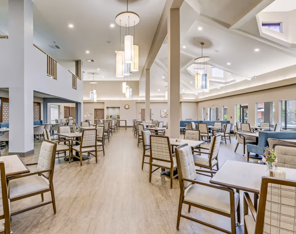 Spacious and well-lit dining room with multiple tables and cushioned chairs arranged neatly. The room features high ceilings with modern hanging light fixtures and large windows allowing natural light to enter. The flooring is wooden, and the overall decor is neutral and inviting.