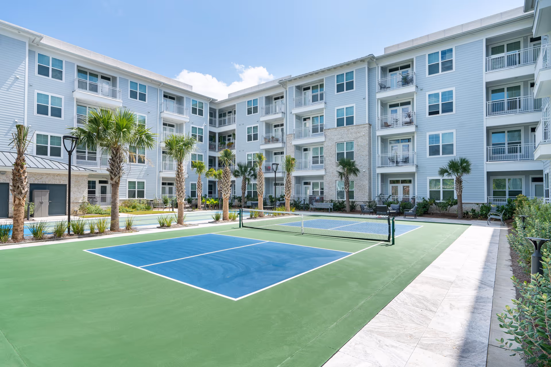 Outdoor courtyard area of a senior living facility with a pickleball court surrounded by palm trees and a four-story building with balconies and large windows under a partly cloudy sky.