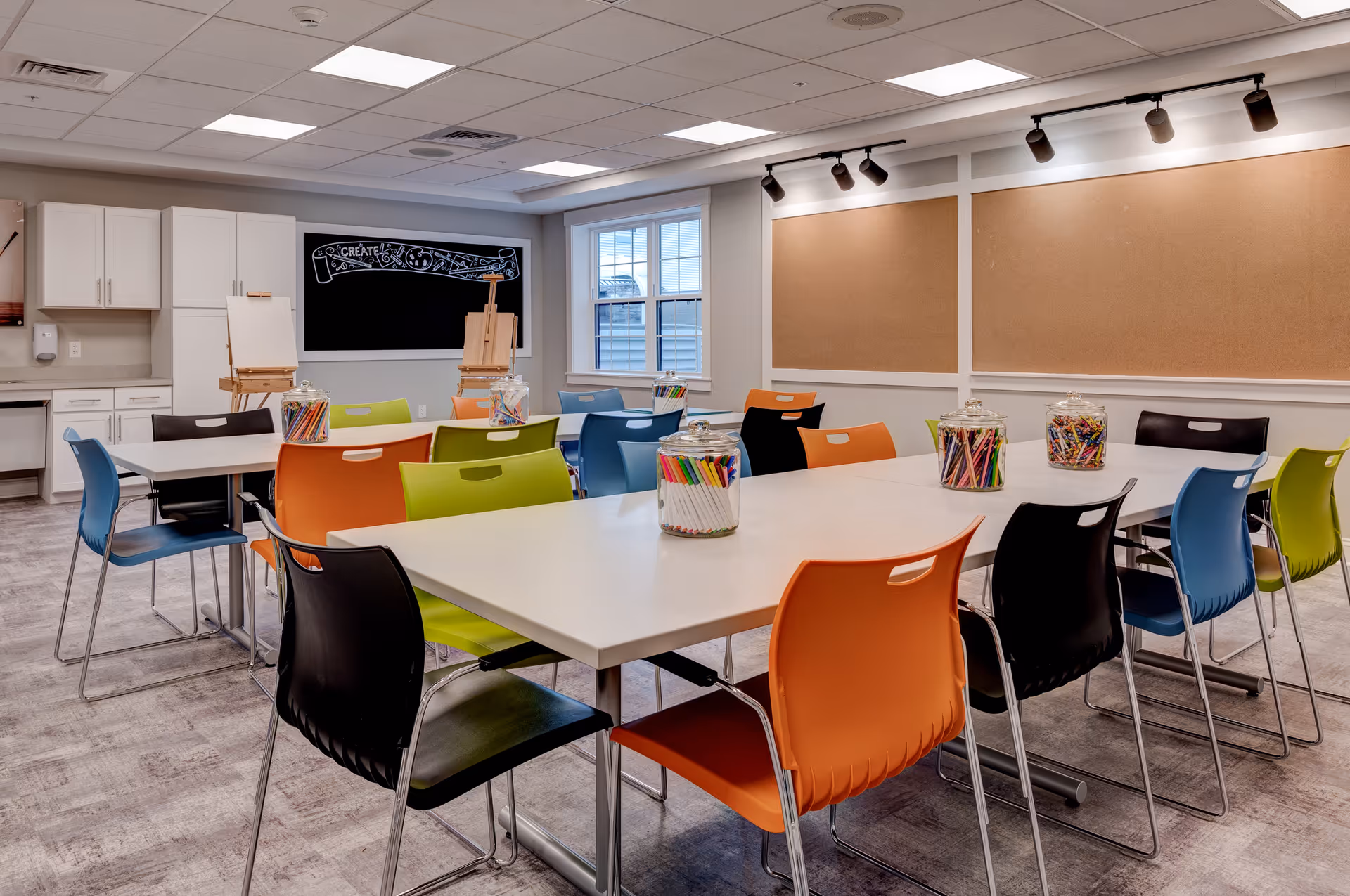A bright and colorful activity room with multiple tables and chairs in orange, green, blue, and black. Each table has jars filled with colored pencils and markers. There are two easels with blank canvases near a blackboard that has the word 'CREATE' written on it. The room has white cabinets, a window, and cork bulletin boards on the wall.