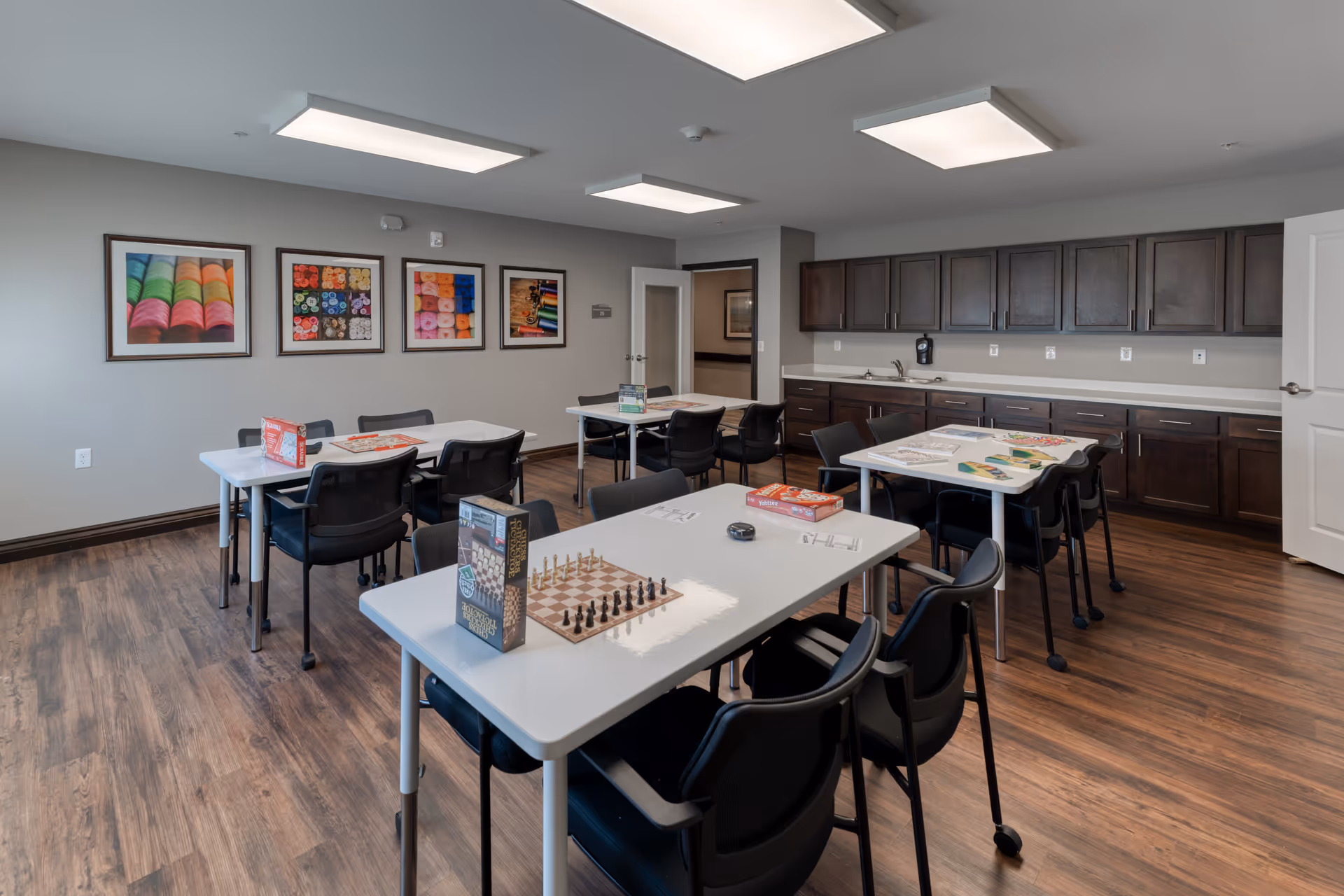 A well-lit activity room with several white tables and black chairs arranged around them. On the tables are various board games and puzzles, including chess and Yahtzee. The room has wooden flooring, gray walls adorned with colorful framed pictures, and dark wooden cabinets along one wall with a countertop and sink. A door is open leading to another room.