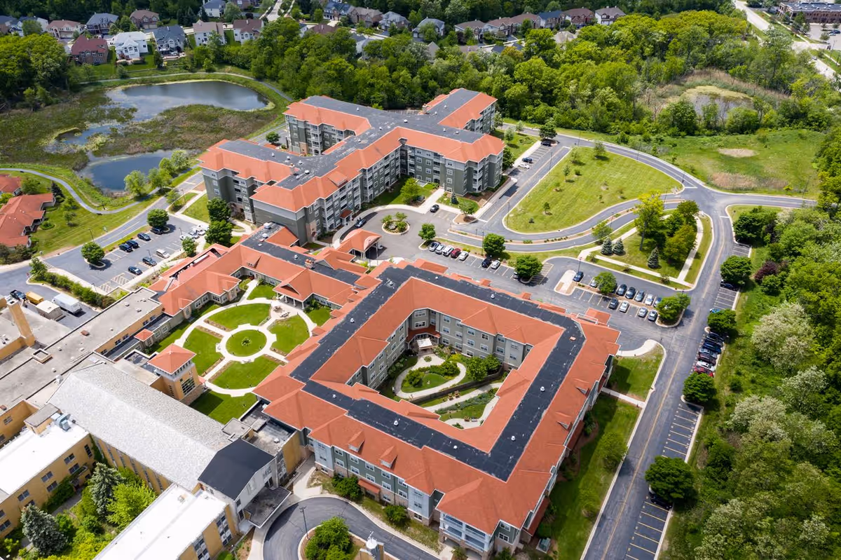Aerial view of The Oaks at Bartlett senior living facility showing multiple connected buildings with red roofs, landscaped courtyards, parking lots, surrounding greenery, and nearby residential houses.