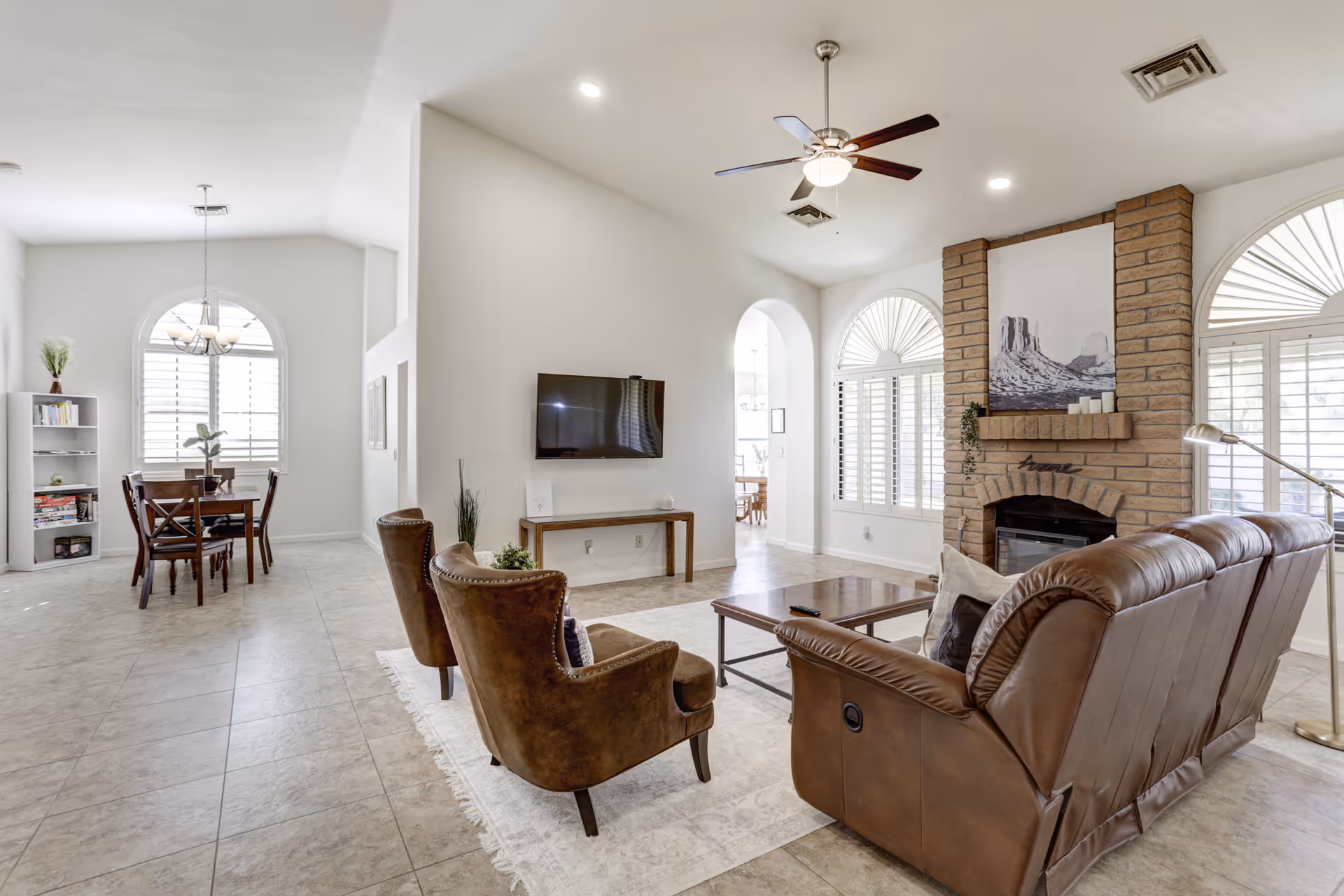 A bright and spacious living room area with a brown leather sofa and two brown armchairs arranged around a wooden coffee table on a light rug. The room features a brick fireplace with candles and a black and white picture above it. There is a wall-mounted flat-screen TV opposite the seating area. Large arched windows with white shutters allow natural light to fill the space. In the background, a dining area with a wooden table and chairs is visible, along with a bookshelf and a chandelier.