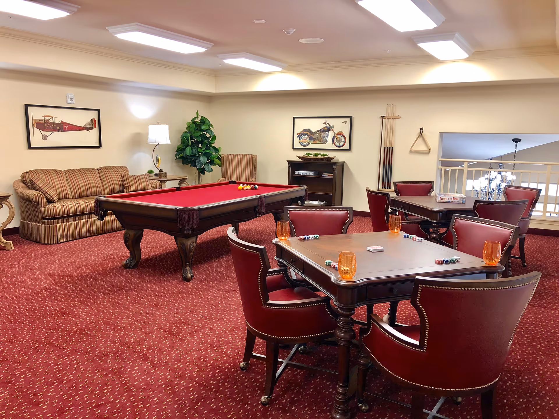 A cozy game room with a red-felt pool table in the center, surrounded by a striped sofa, a tall green plant, and framed pictures of a vintage airplane and a motorcycle on the walls. There are two wooden tables with red leather chairs arranged around them, each table set with poker chips, playing cards, and orange drinking glasses. The room has a red carpet with a subtle pattern and bright ceiling lights.