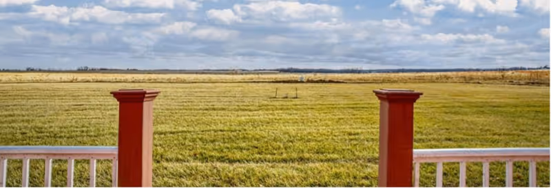 View over a wide grassy field from a porch with white railing and two red posts under a cloudy sky.