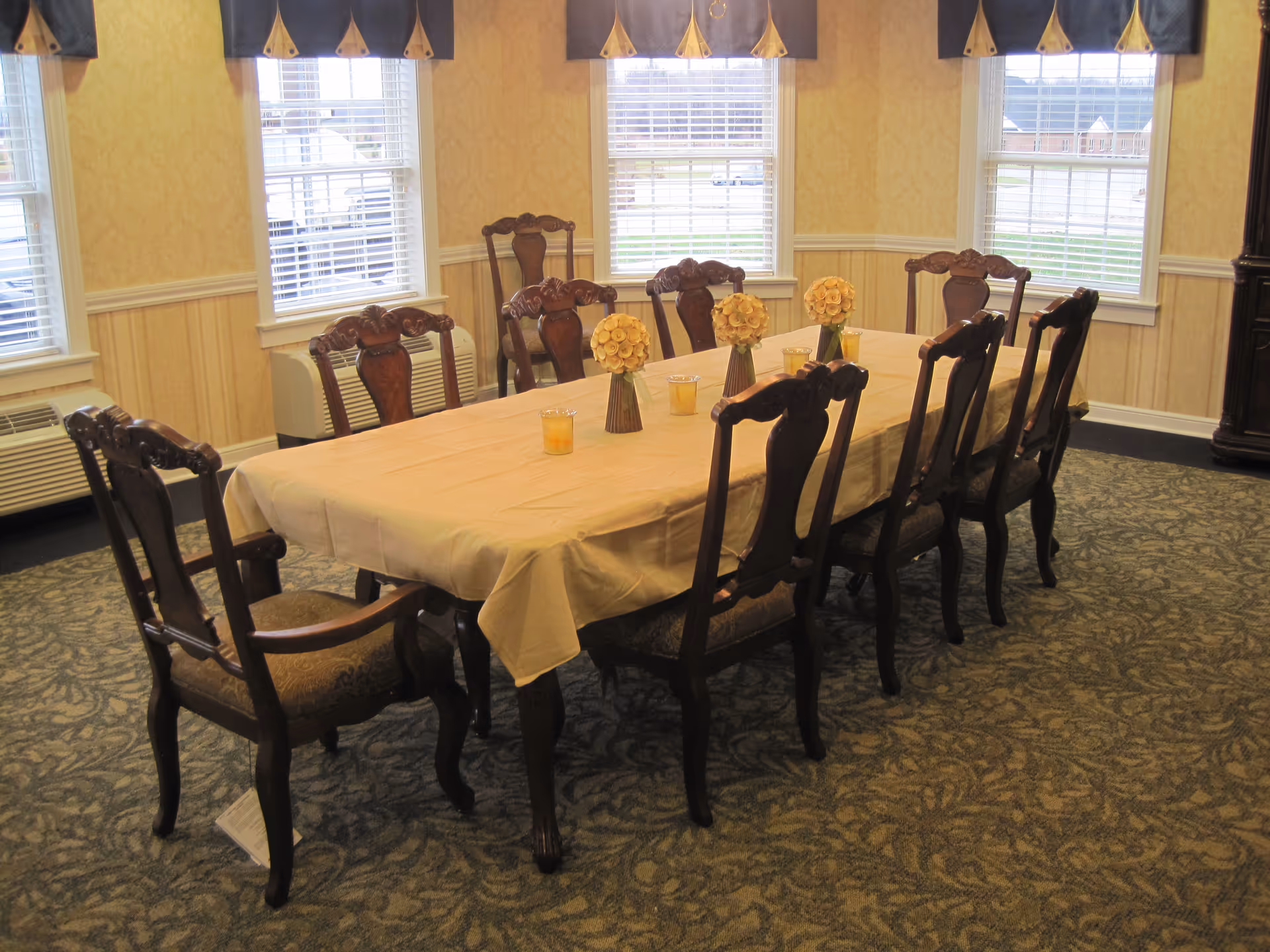 A dining room with a long rectangular table covered with a beige tablecloth, surrounded by ten wooden chairs with cushioned seats. The table is decorated with four small flower arrangements and four candles. The room has patterned carpet, yellow wallpaper, and four windows with white blinds and dark valances.