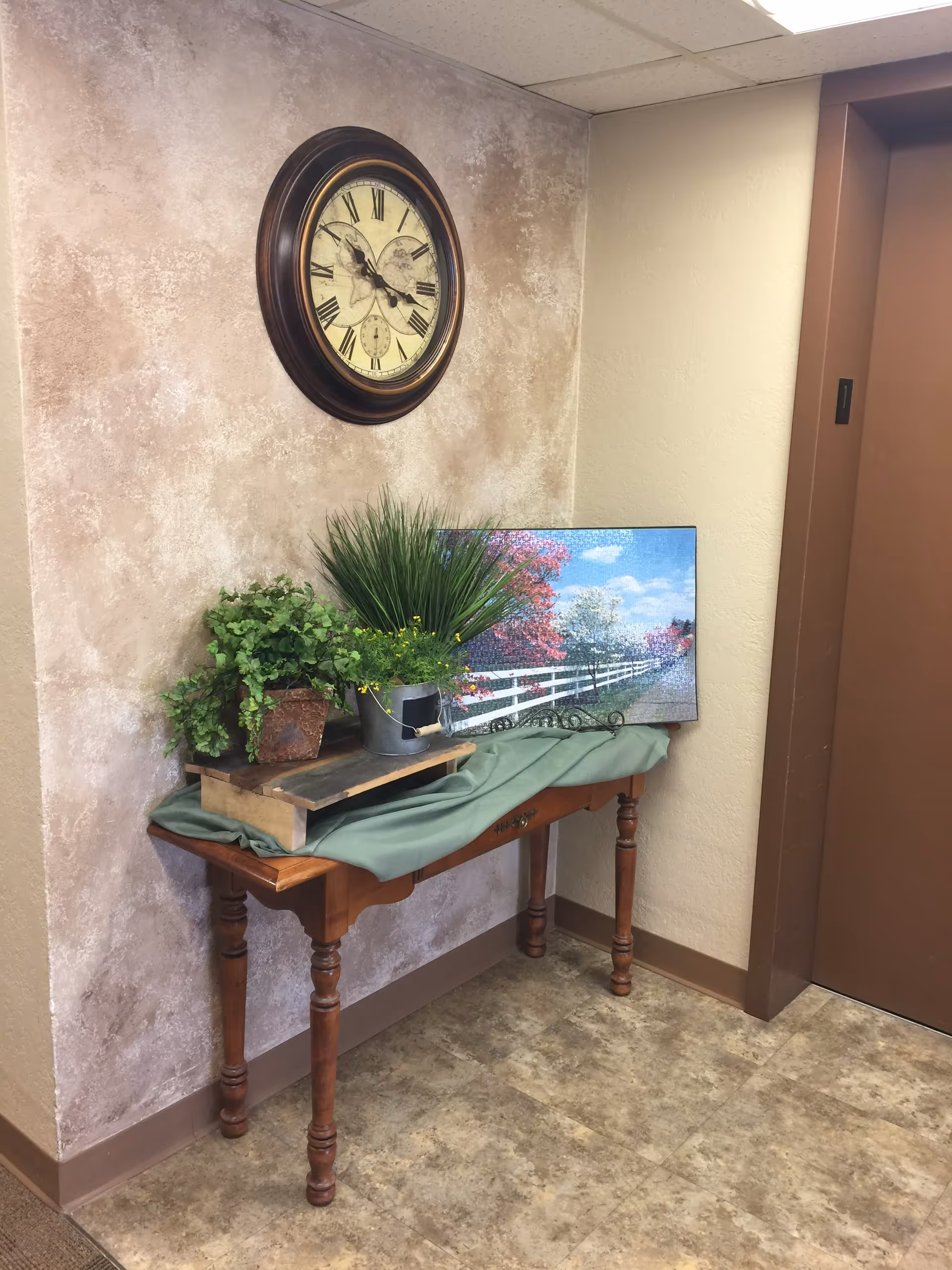 A corner of an interior space with a wooden table against a textured wall. On the table are two potted plants, a green cloth, and a framed picture of a white fence with trees and a blue sky. Above the table is a round wall clock with Roman numerals. To the right is a brown elevator door with the number 1 on it.