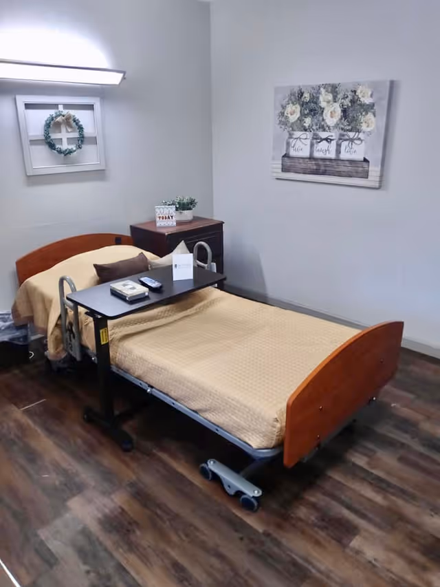 A single hospital-style bed with wooden headboard and footboard in a nursing center room. The bed is covered with a beige blanket and has a small brown pillow. A black overbed table holds a book, a remote control, and a folded card. Next to the bed is a small wooden nightstand with a potted plant and a decorative sign. The walls are light gray with two pieces of wall art: a small wreath in a white frame and a floral painting with the words 'live', 'laugh', and 'love'. The floor is dark wood laminate.