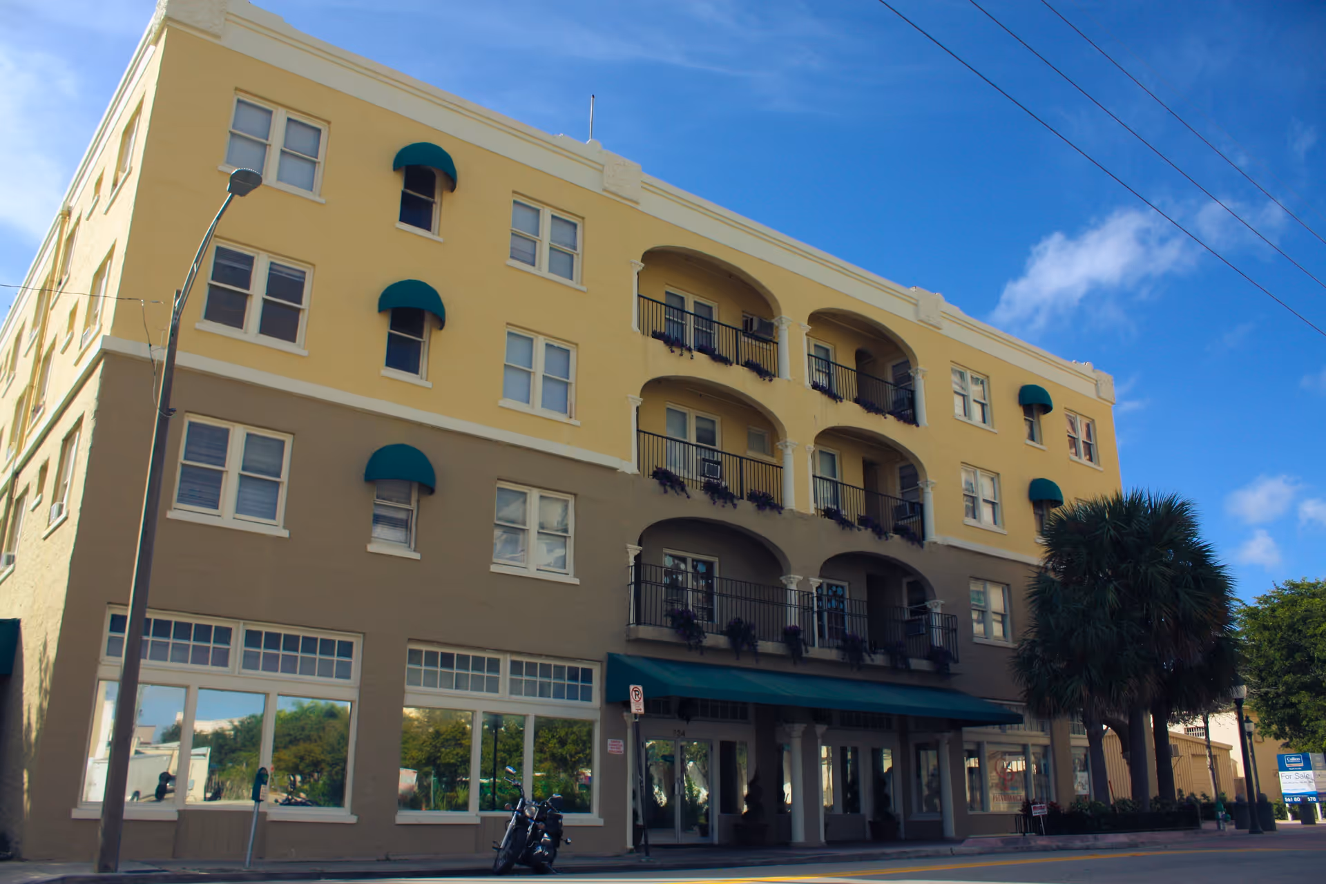 Exterior view of a multi-story building with a yellow and beige facade, featuring balconies with black railings and purple flowers. There is a green awning above the entrance, a palm tree on the right side, and a motorcycle parked on the street in front. The sky is clear and blue with some clouds.