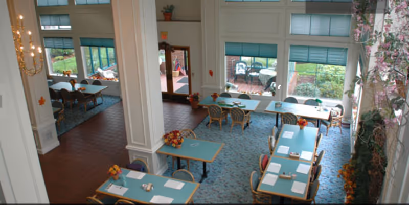 Interior view of a dining room with multiple tables and chairs arranged neatly. The room has large windows allowing natural light to brighten the space, with a view of an outdoor patio area. The tables have small flower arrangements and papers placed on them. The floor is carpeted with a patterned design, and there are white pillars and decorative elements around the room.