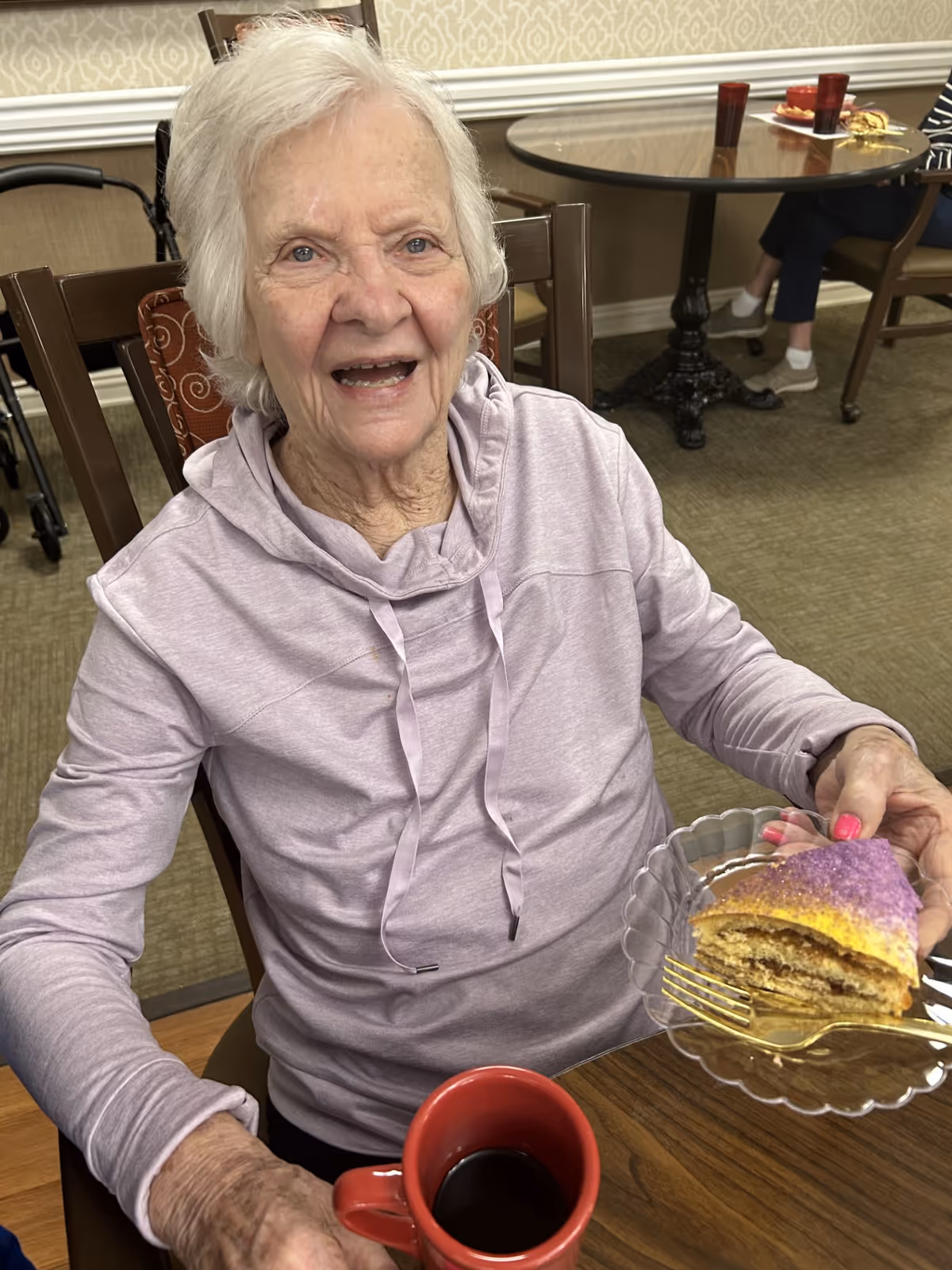 An elderly woman with white hair wearing a light purple hoodie is sitting at a wooden table in a dining area. She is smiling and holding a clear plate with a slice of layered cake topped with purple and yellow sugar. In front of her is a red mug filled with a dark beverage. In the background, another person is seated at a round table with red cups and plates.