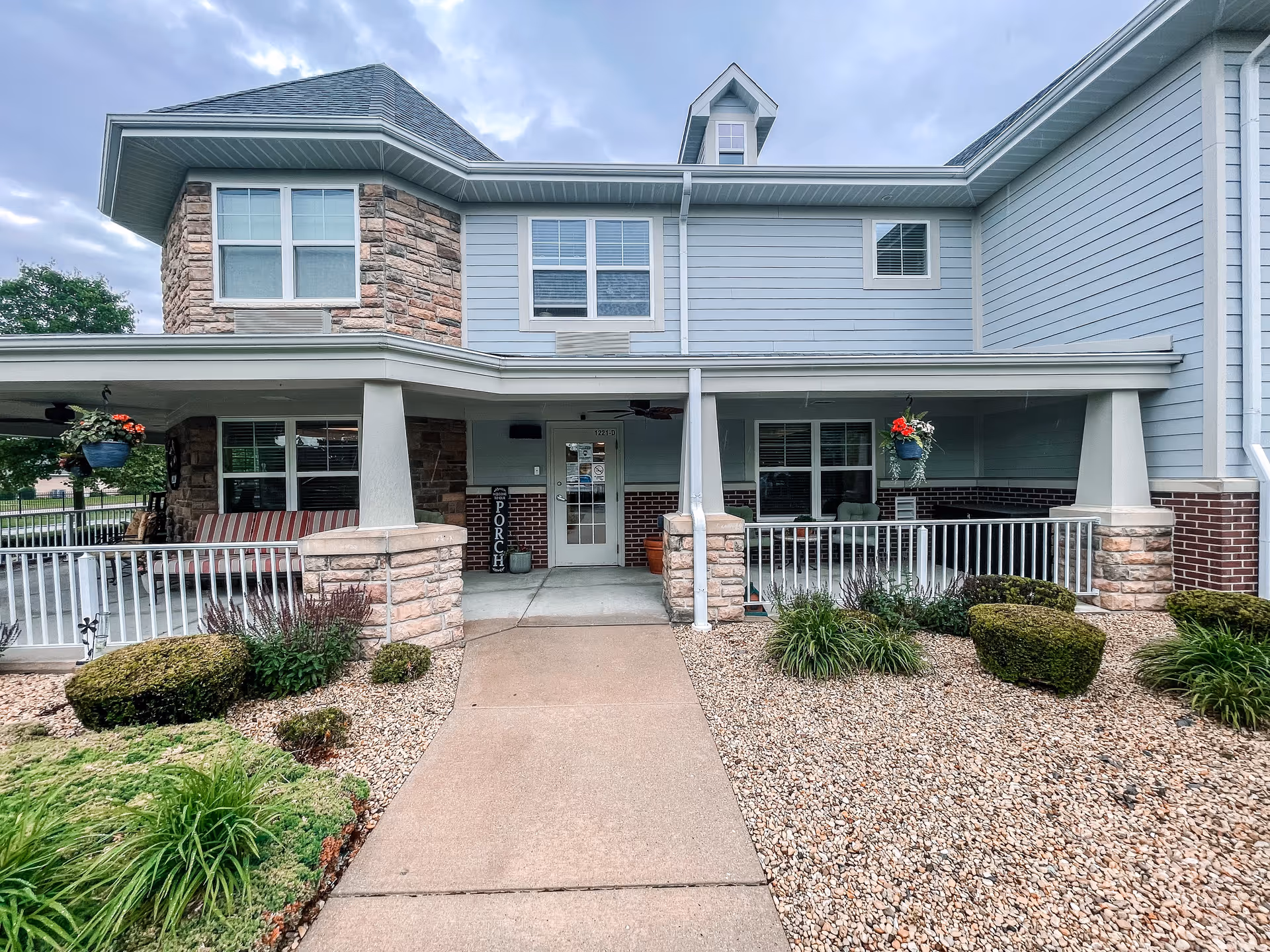 Front exterior view of a two-story building with a covered porch area. The building features a combination of stone and light blue siding with white trim. There are hanging flower pots on the porch, neatly trimmed bushes, and a concrete walkway leading to the entrance door labeled 'PORCH'.