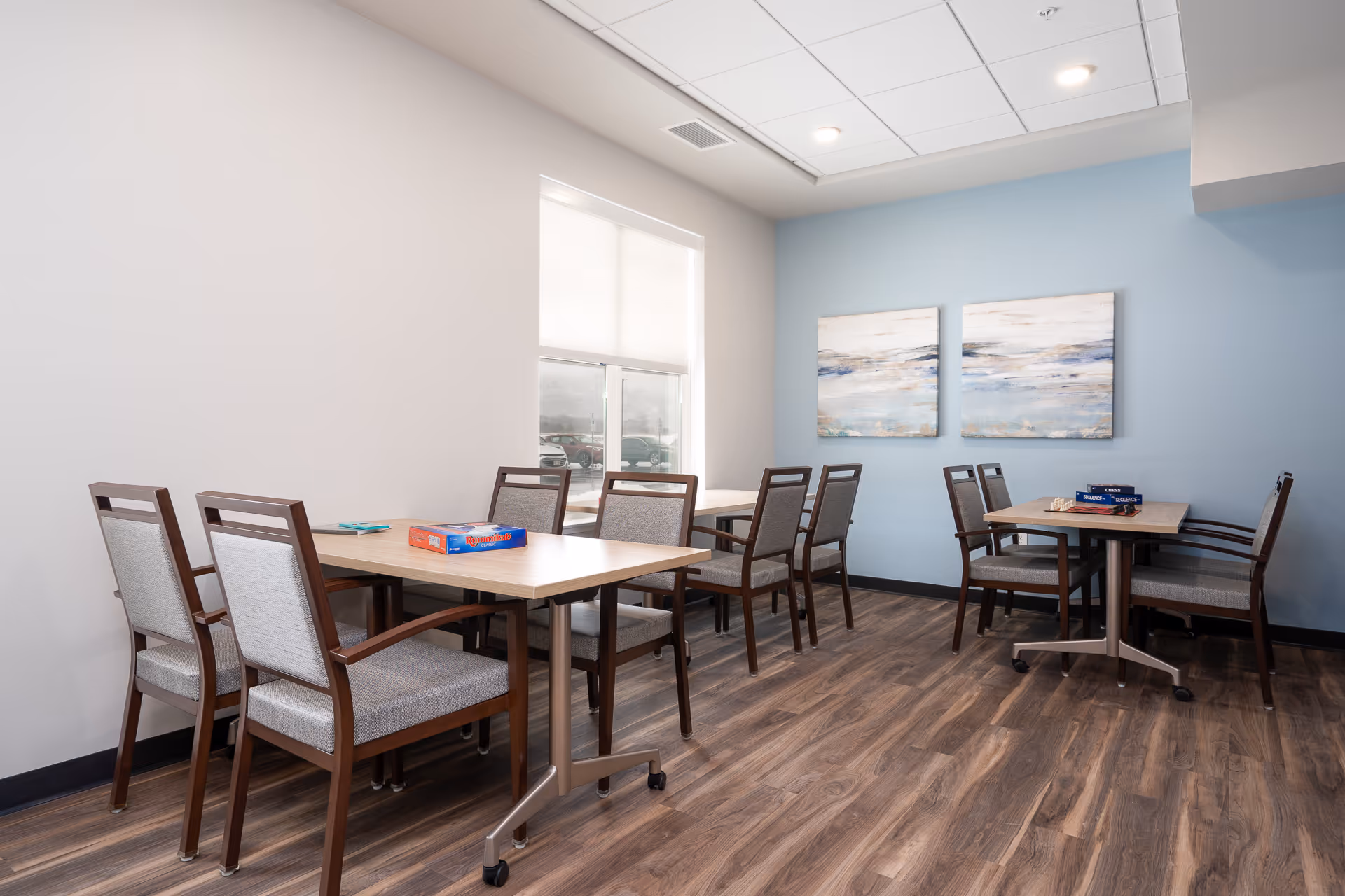 Bright senior living dining/activity room with multiple tables and chairs, wood flooring, a blue accent wall and two abstract paintings.