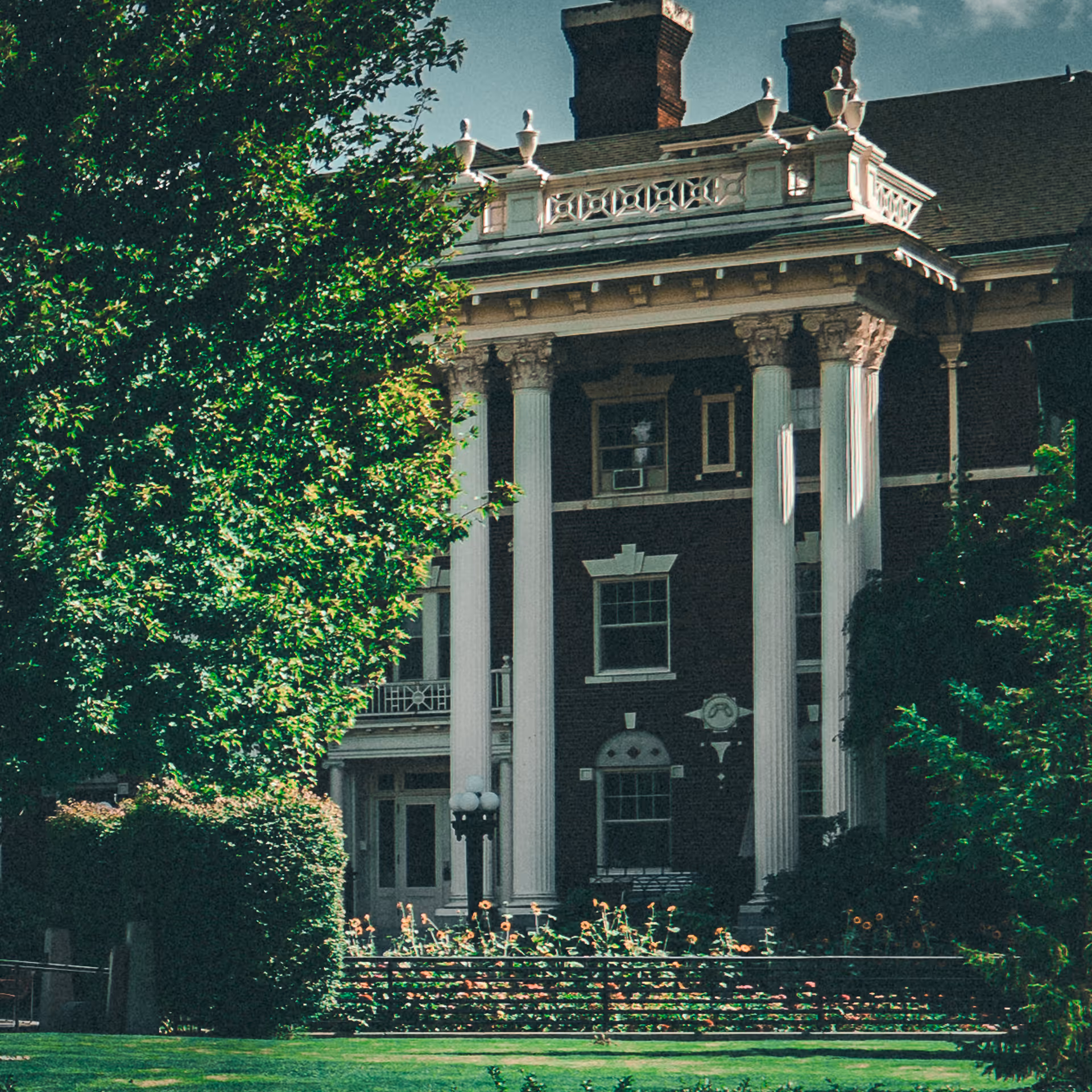 Exterior view of a large historic brick building with tall white columns and decorative architectural details, partially obscured by green trees and bushes, with a well-maintained lawn and flower bed in front.