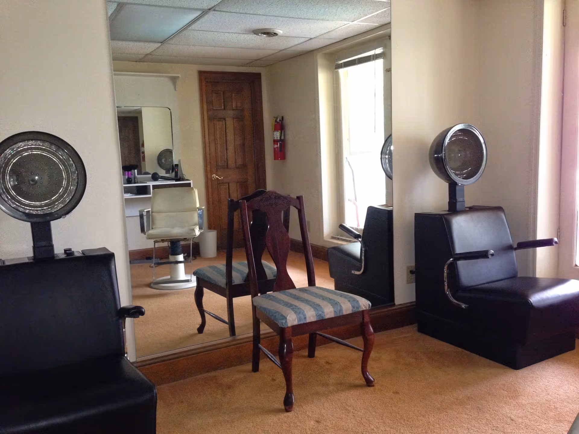 Interior of a hair salon area with two black vintage hair dryer chairs, a wooden chair with striped upholstery, a large wall mirror, a window, and a wooden door in the background.