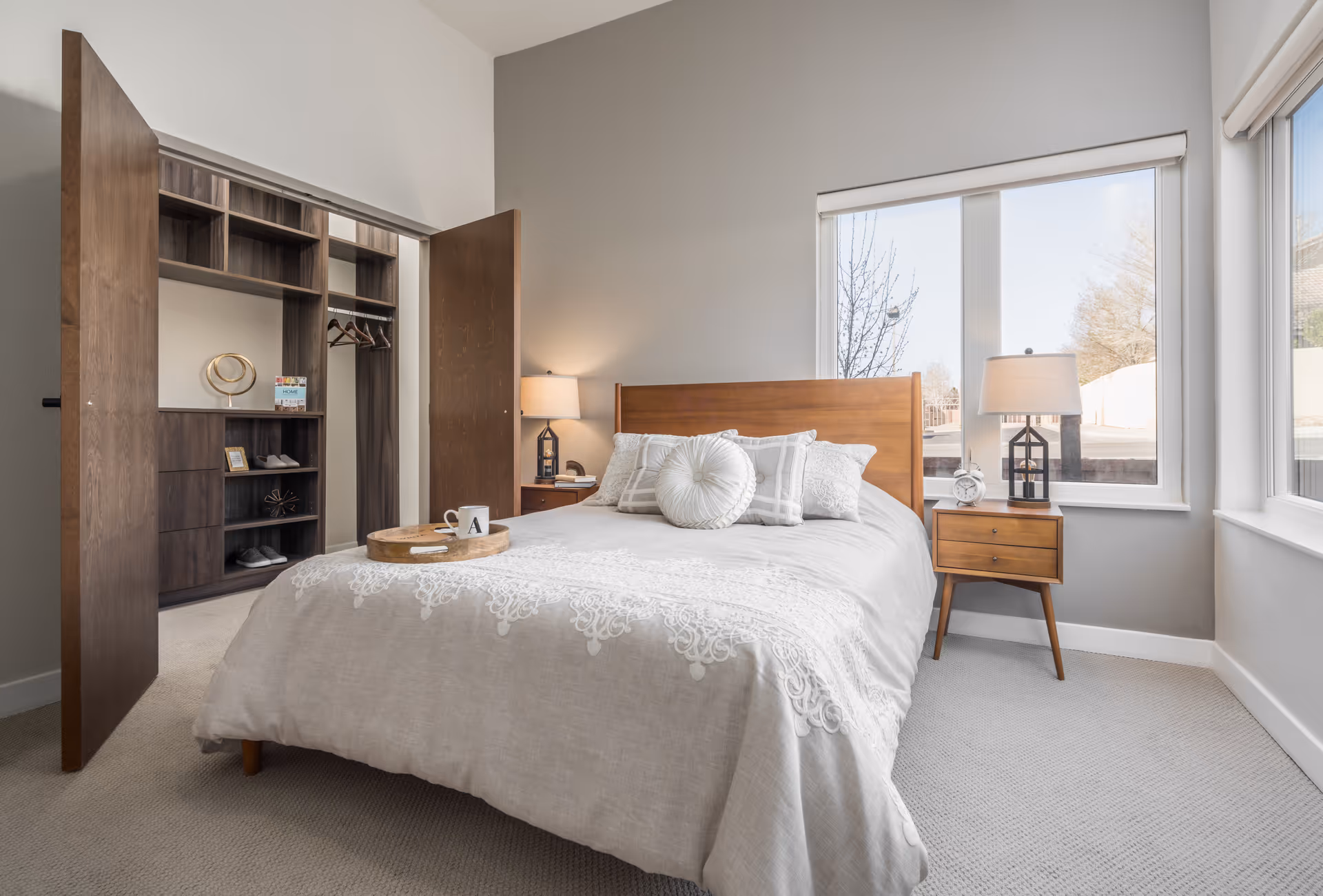 A bright bedroom with a wooden bed frame and a beige bedspread adorned with decorative pillows. There is a wooden nightstand with a lamp and a clock next to the bed. The room has large windows letting in natural light and an open closet with wooden shelves and hanging space.