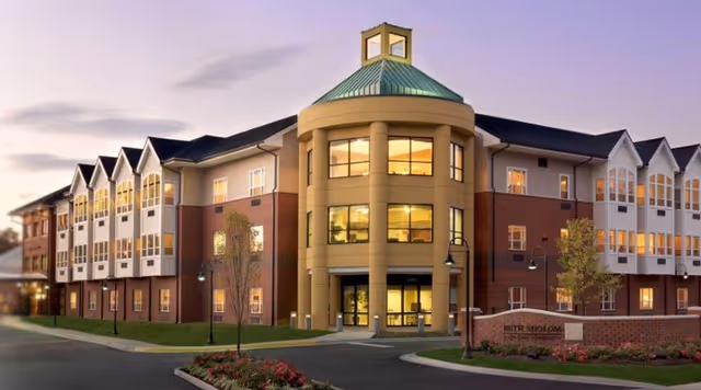 Exterior view of a large, modern senior assisted living facility building at dusk with multiple windows lit from inside, a central rounded entrance with columns, a green roof cupola, landscaped grounds, and a sign reading 'Beth Sholom'.