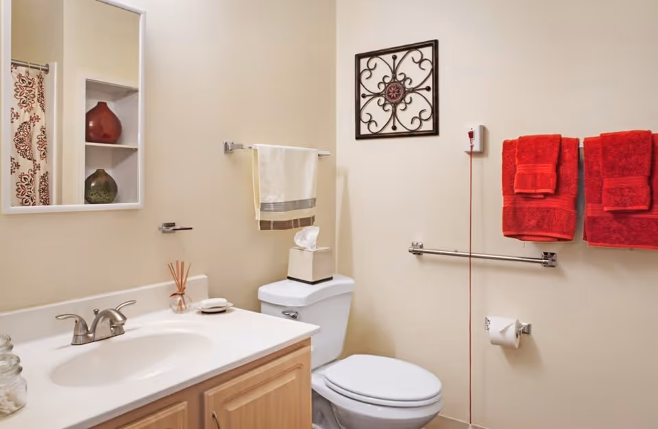 A clean bathroom with a white sink and countertop, a wooden cabinet below, a toilet with a tissue box on top, and a towel rack holding white and red towels. There is a decorative wall hanging above the toilet and a mirror reflecting a shower curtain with a red and beige pattern.