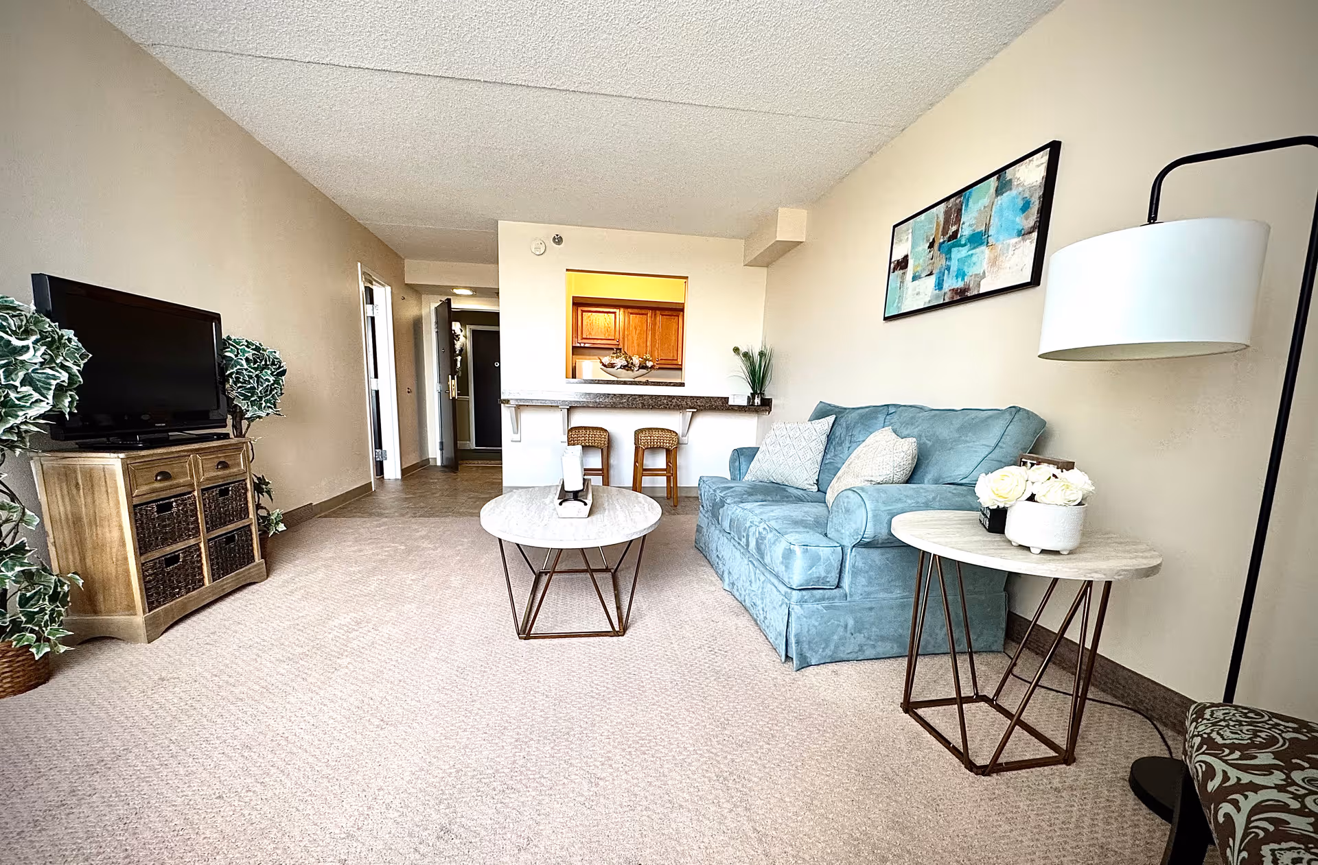 A bright and spacious living room in a senior living facility featuring a light blue sofa with decorative pillows, a round coffee table, a side table with white flowers, a floor lamp, and a TV on a wooden stand with wicker baskets. The room has beige walls and carpet, with a kitchen area visible in the background with two bar stools at a counter.
