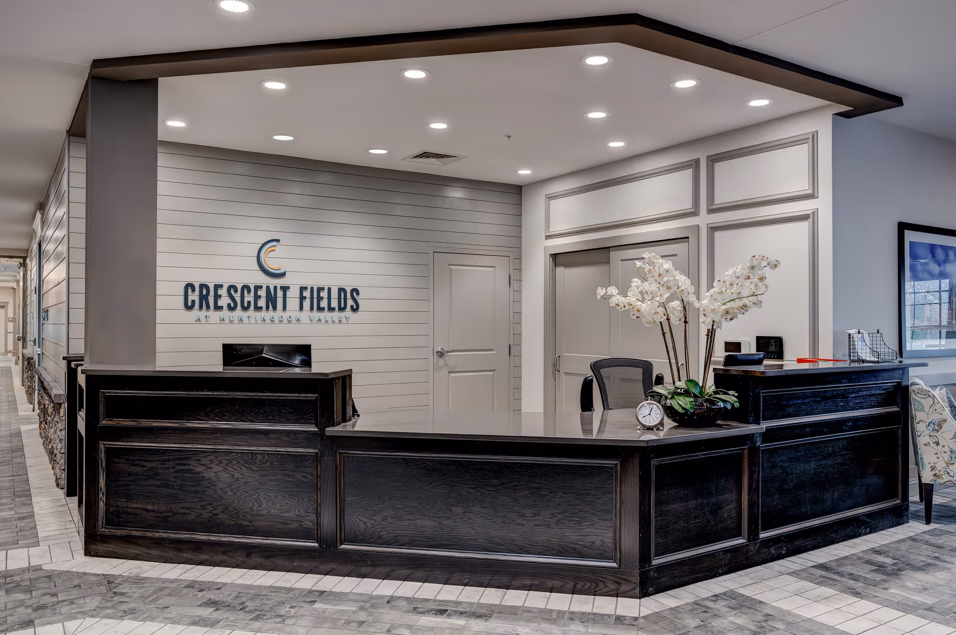 Reception area of Crescent Fields at Huntingdon Valley featuring a dark wood front desk with a vase of white orchids and a small clock on it. Behind the desk is a wall with the facility's name and logo, a door, and a hallway to the left.