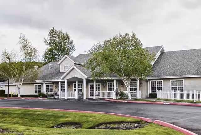 Exterior view of a single-story senior living facility building with a covered entrance, white railings, several windows, and trees around the property under a cloudy sky.