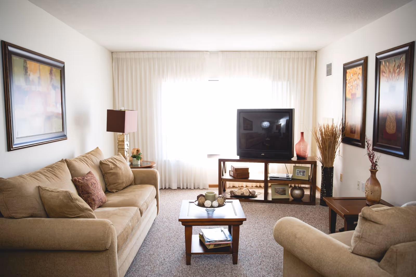 A cozy living room with beige sofas, a wooden coffee table with decorative items, a TV on a wooden stand, and framed artwork on the walls. The room is softly lit by natural light coming through sheer white curtains covering a large window.