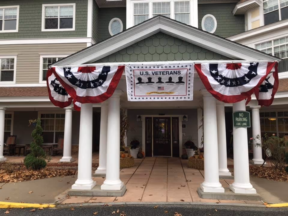 Entrance of a senior living facility decorated with red, white, and blue bunting and a banner honoring U.S. Veterans. The entrance has white columns and a covered walkway leading to glass doors. There is a 'No Parking Fire Lane' sign on one of the columns.