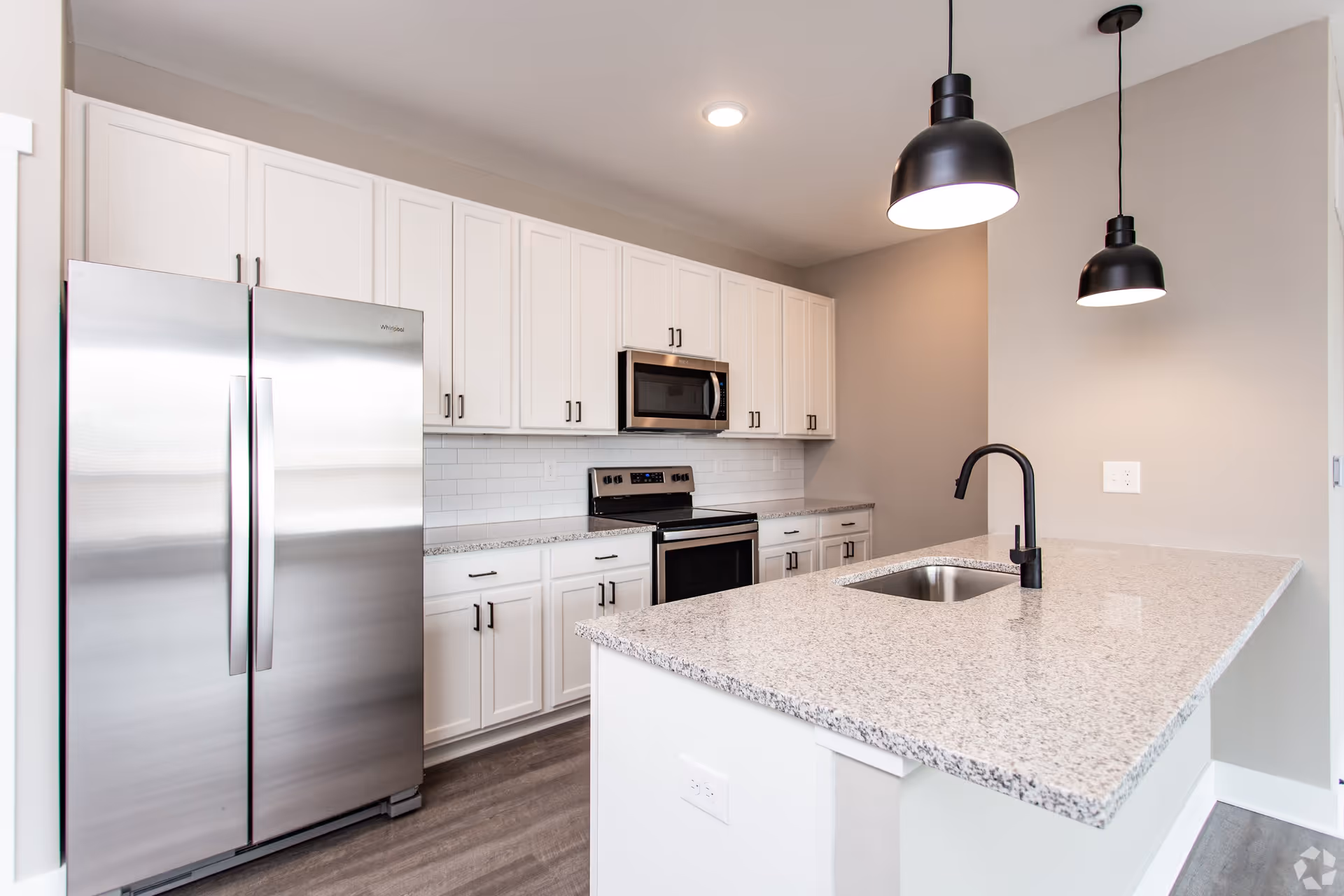 Modern kitchen with white cabinets, stainless steel refrigerator, oven, microwave, and a granite countertop island with a built-in sink and black faucet. Two black pendant lights hang above the island.