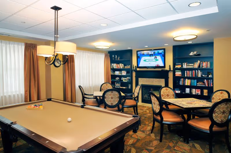A cozy recreational room with a pool table in the foreground, a round table with chairs, bookshelves filled with books, a fireplace, and a wall-mounted TV displaying a curling game. The room has warm lighting, patterned carpet, and large windows with curtains.