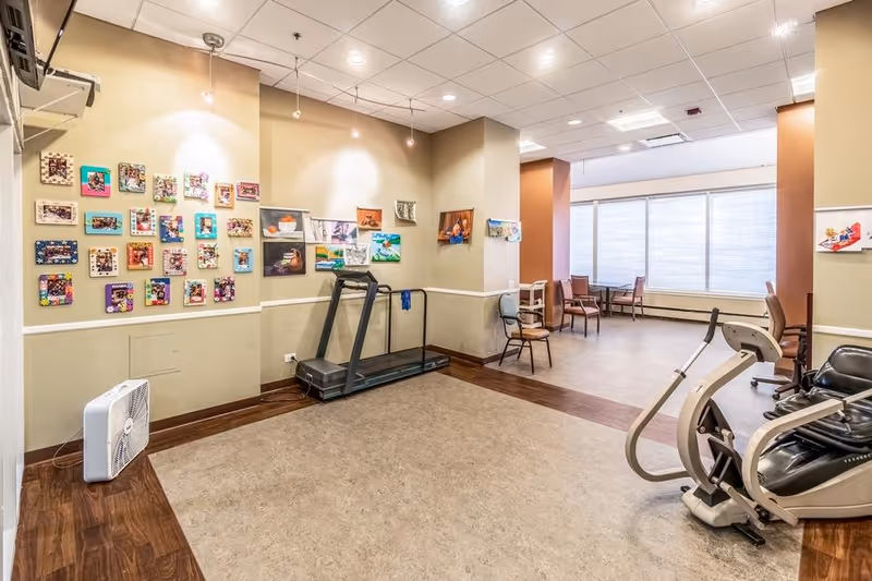 Interior view of a senior living facility room with exercise equipment including a treadmill and a stationary bike. The walls are decorated with colorful artwork and photos. There are several chairs and tables near large windows with blinds, allowing natural light to fill the space.