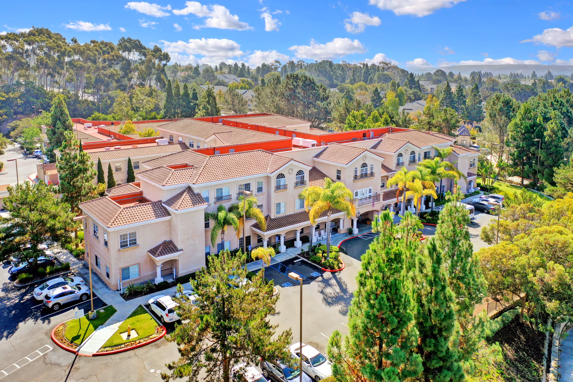 Aerial view of a large senior living facility building with beige walls and red-tiled roofs surrounded by trees and greenery under a blue sky with scattered clouds. Several cars are parked in the parking lot around the building.