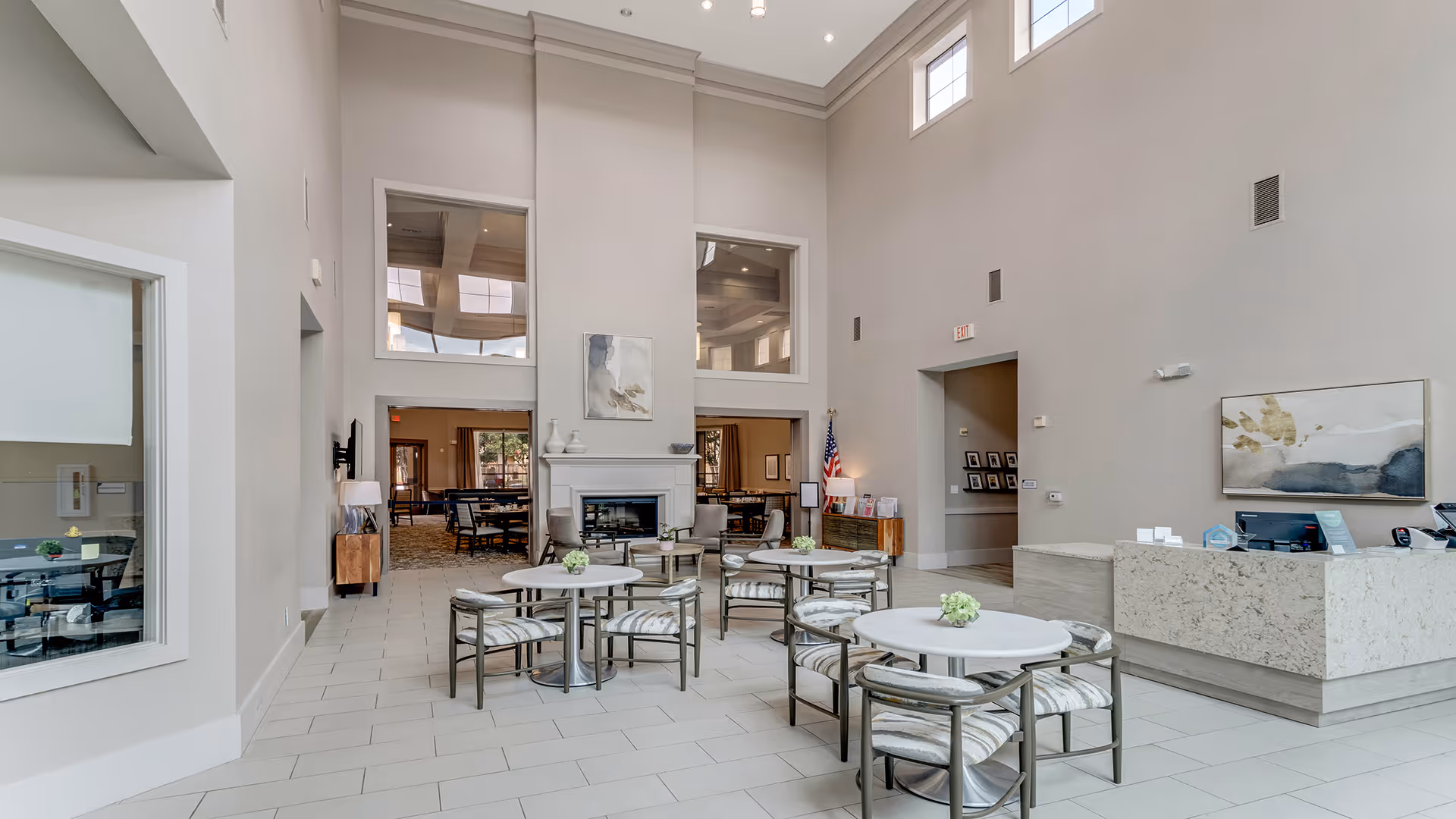 A spacious and bright common area in a senior living facility with high ceilings and large windows. The room features several round tables with chairs arranged around them, each table decorated with a small floral centerpiece. There is a modern fireplace in the center of the back wall, flanked by two large windows. To the right, there is a reception desk with a computer and office supplies. The walls are painted light gray, and the floor is tiled in a light color. An American flag stands near the back wall, and there is abstract artwork hanging on the walls.
