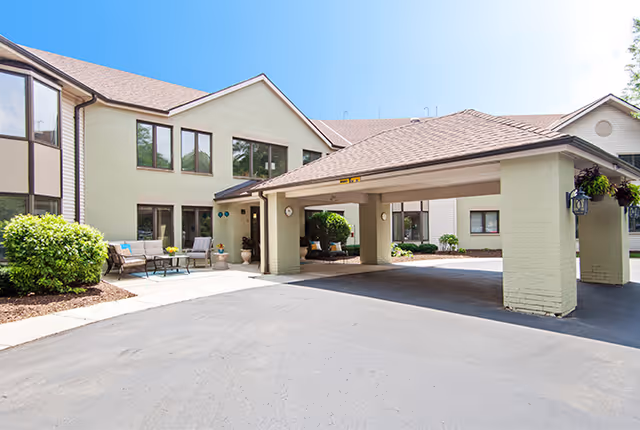 Exterior view of LakeHouse Menomonee facility showing a two-story building with beige walls and a covered entrance driveway. There are outdoor seating areas with chairs and tables near the entrance, along with some greenery and bushes. The sky is clear and blue.