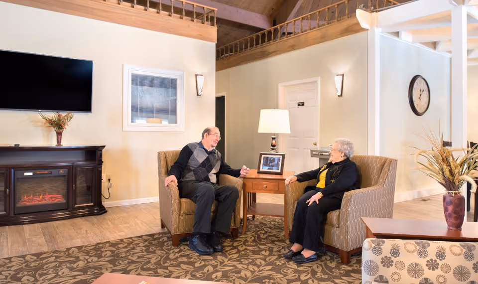 Two elderly people sitting and talking in a comfortable living room area with armchairs, a patterned carpet, a wooden side table with a lamp and framed photo, a wall-mounted TV above a fireplace, and decorative plants.