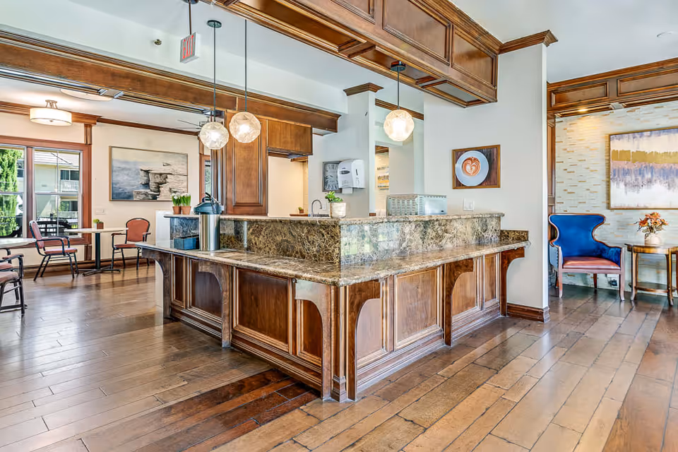 Interior view of a senior living facility with a large wooden reception or service counter featuring a marble countertop. The area has wooden flooring and ceiling accents, pendant lights hanging above the counter, and seating areas with chairs and tables visible in the background. Artwork and plants decorate the walls and surfaces.