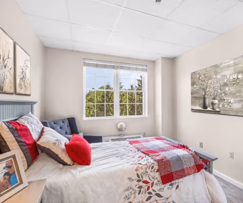Sunlit bedroom with a bed dressed in floral bedding and red accents, a tufted chair by the window, and wall art.