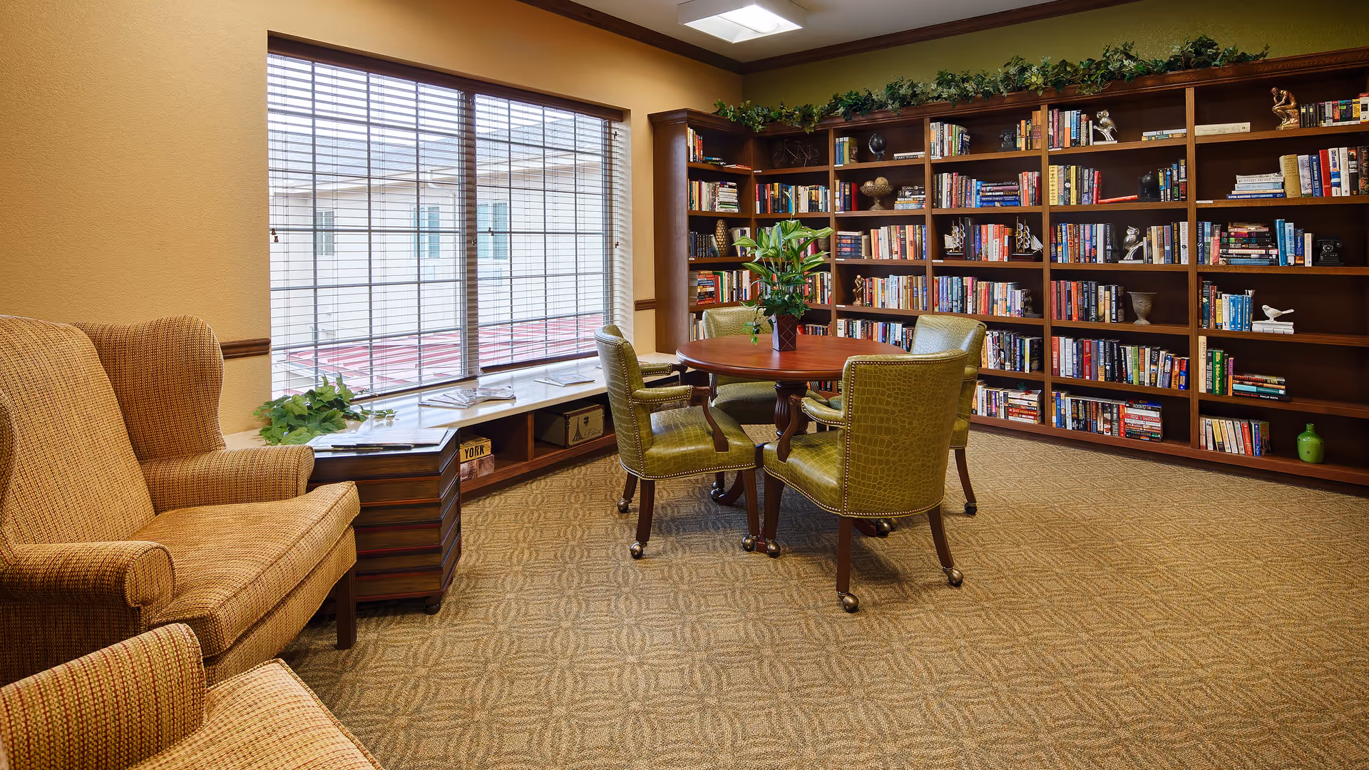 A cozy library room with a large window covered by blinds, a round wooden table surrounded by four green upholstered chairs, a beige armchair, and a wall filled with bookshelves stocked with books and decorative items.