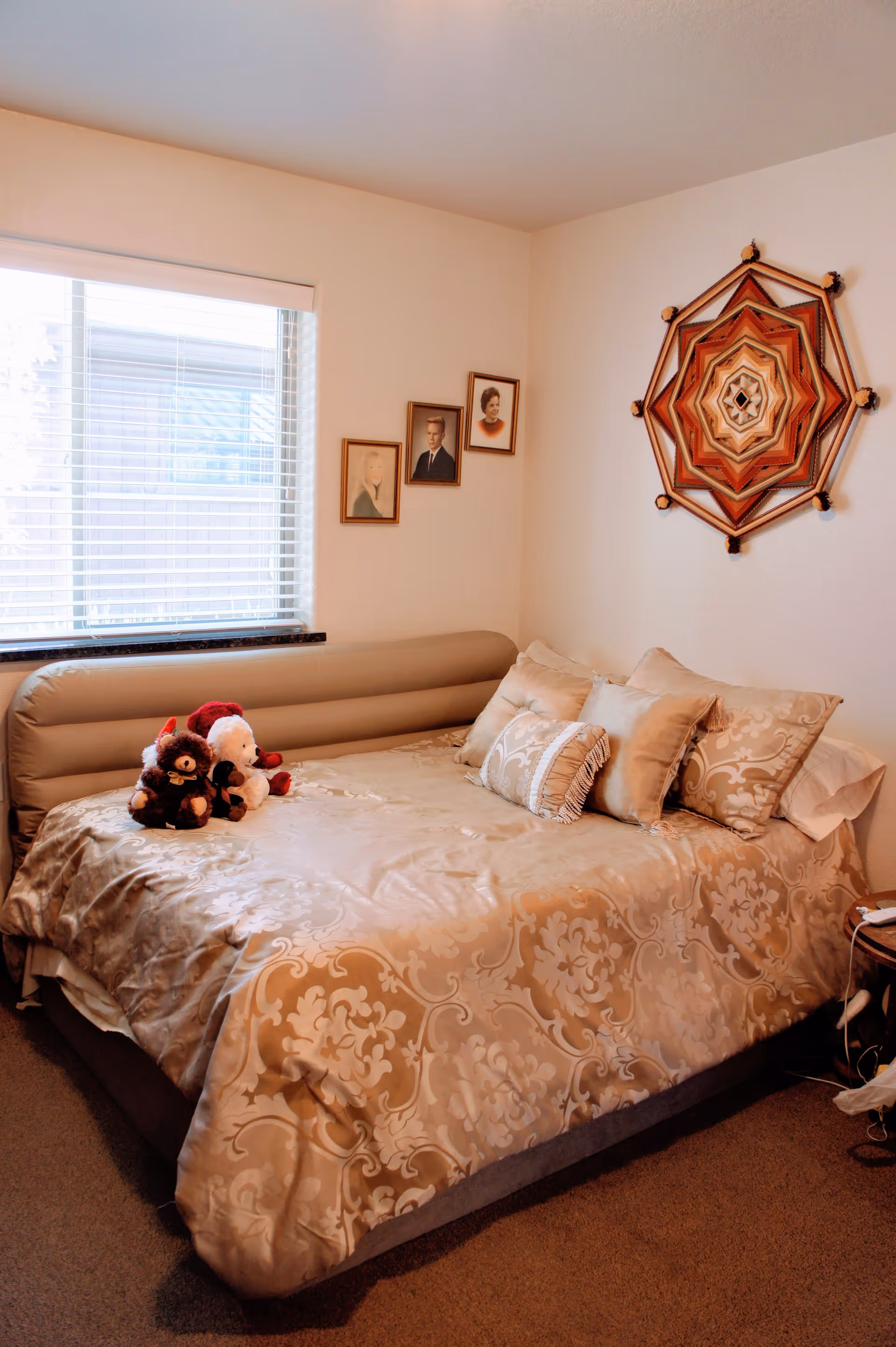 A cozy bedroom with a large bed covered in a beige patterned comforter and several decorative pillows. Three stuffed animals are placed on the bed near the headboard. The room has a window with white blinds on the left wall, and three framed portraits hanging on the wall above the bed. A large, decorative woven wall hanging is displayed on the right wall. The floor is carpeted, and a small round wooden side table is partially visible on the right side.