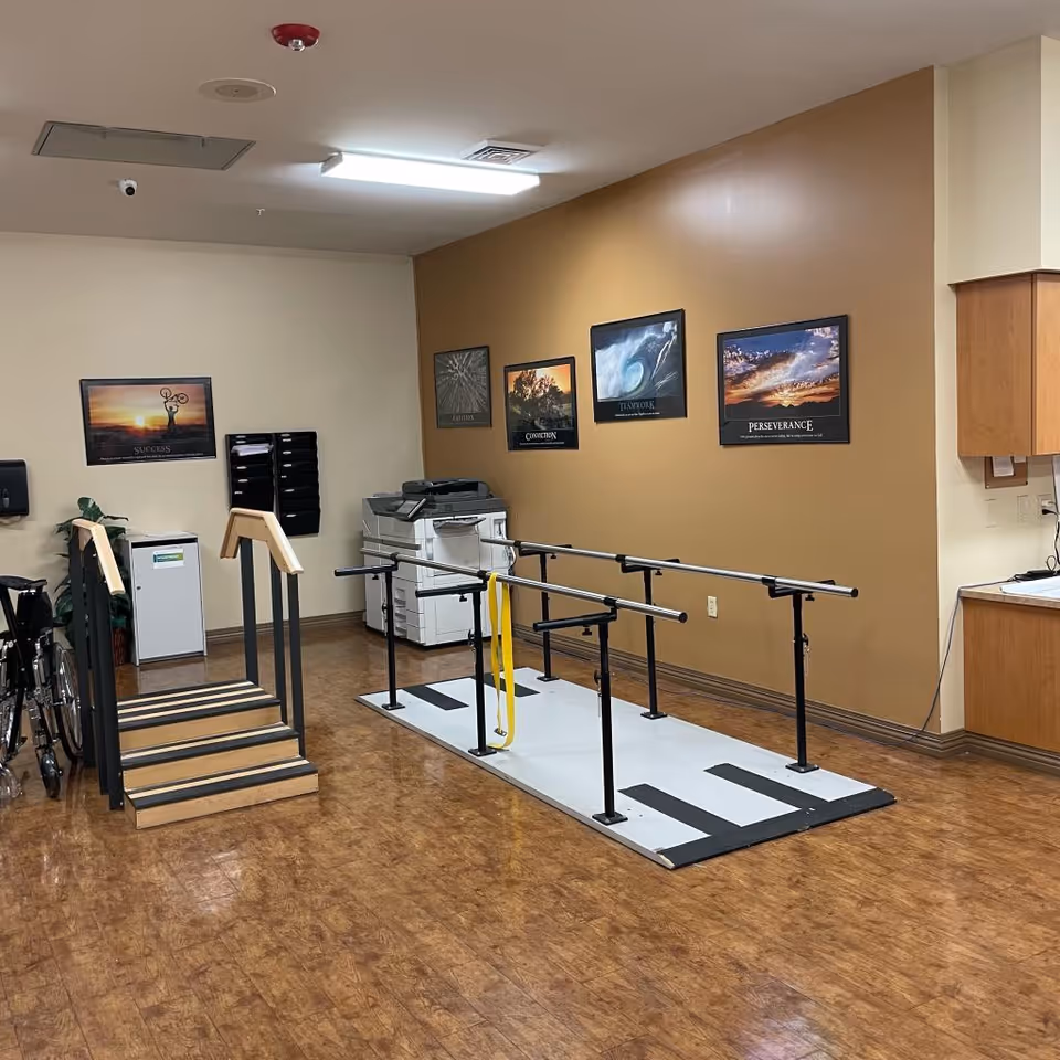 An indoor rehabilitation room with wooden flooring, featuring parallel bars for walking exercises and a small set of stairs with handrails. There is a wheelchair on the left side, a printer and filing trays against the wall, and motivational posters on the walls. A countertop with cabinets is visible on the right side.