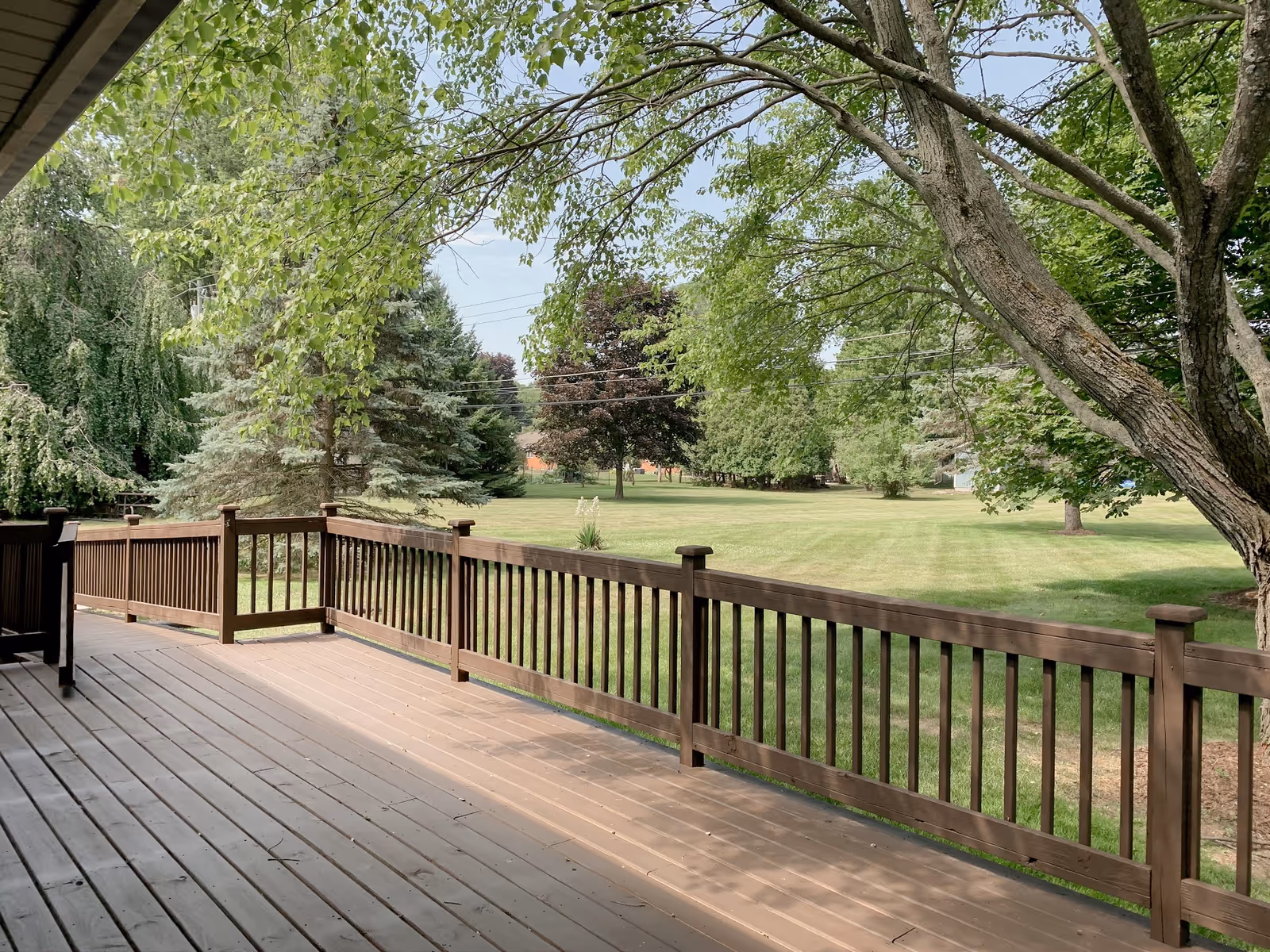 A wooden deck with railing overlooking a large grassy yard with several trees and a clear sky.